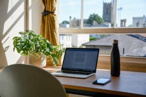 A desk with a green plant, computer, phone, and water bottle on it, looking out a window with a city view